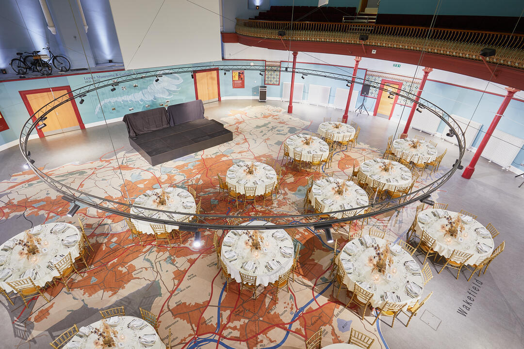 Banquet tables set for an event in a large hall with a regional map design on the floor and a small stage at one end.