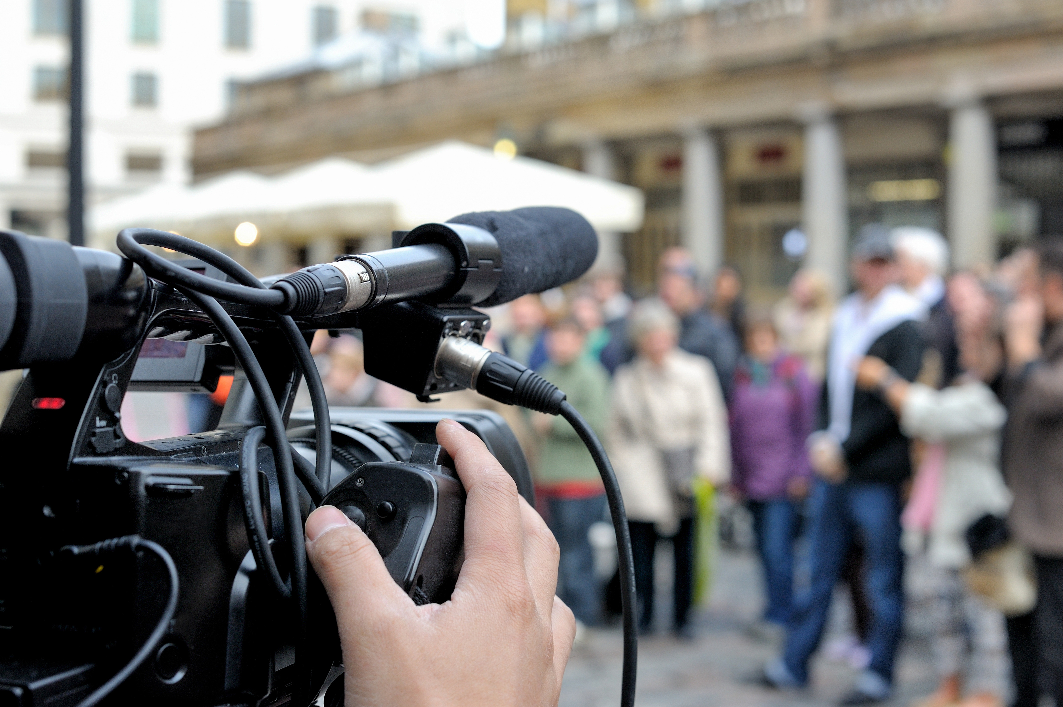 Nahaufnahme der Rückseite einer Fernsehkamera mit einem externen Richtmikrofon und einem an der Kamera befestigten XLR-Kabel. Die Hand des Kameramanns auf dem Fokusgriff, im Hintergrund unscharf abgebildete Personen.