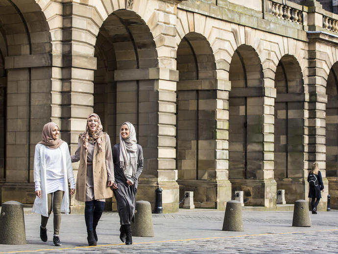 Three women walking by stone arches in a city