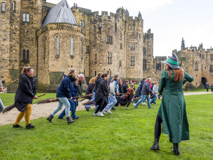 A group of people lined up for a Broomstick Training lesson with the wizarding professors by a large castle.