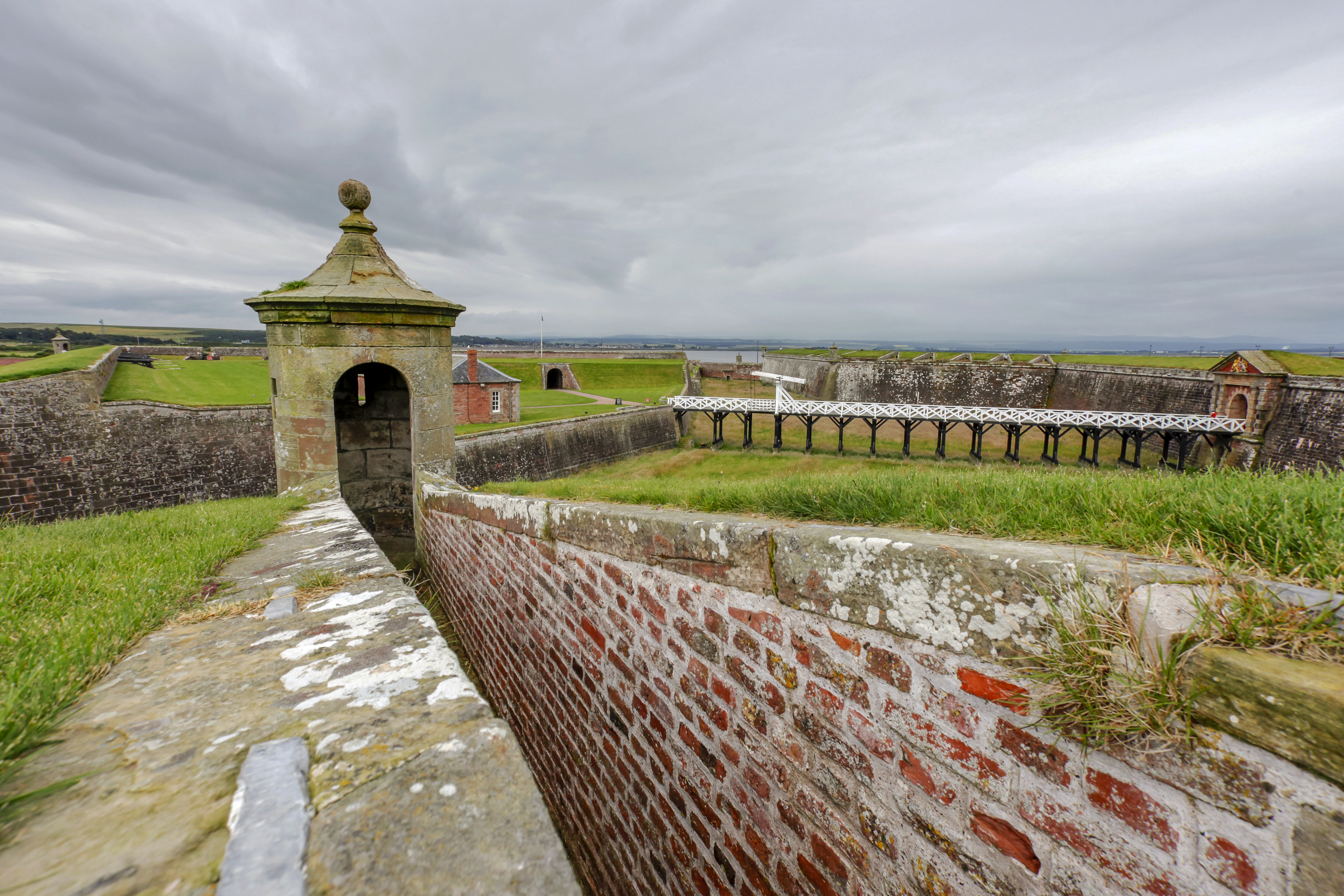 A brick defensive wall with a small tower amid a larger stone fortification under grey skies.
