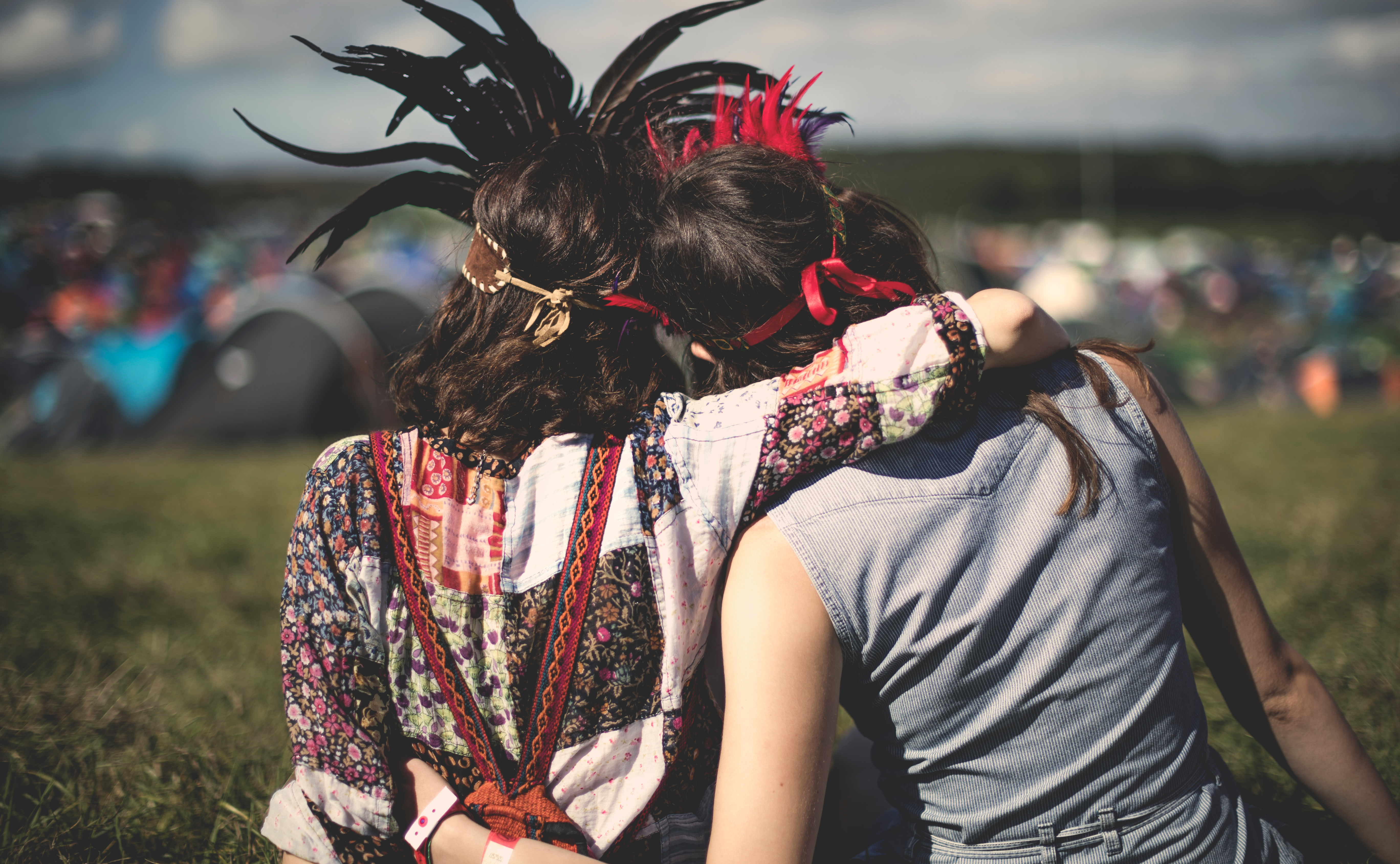 Two people in colorful outfits and feather headbands sit on grass at an outdoor festival, arms around each other, tents in the background.