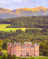 Extérieur d'un palais en grès avec des tourelles ornées, entouré de bois avec de hautes montagnes au loin en automne.