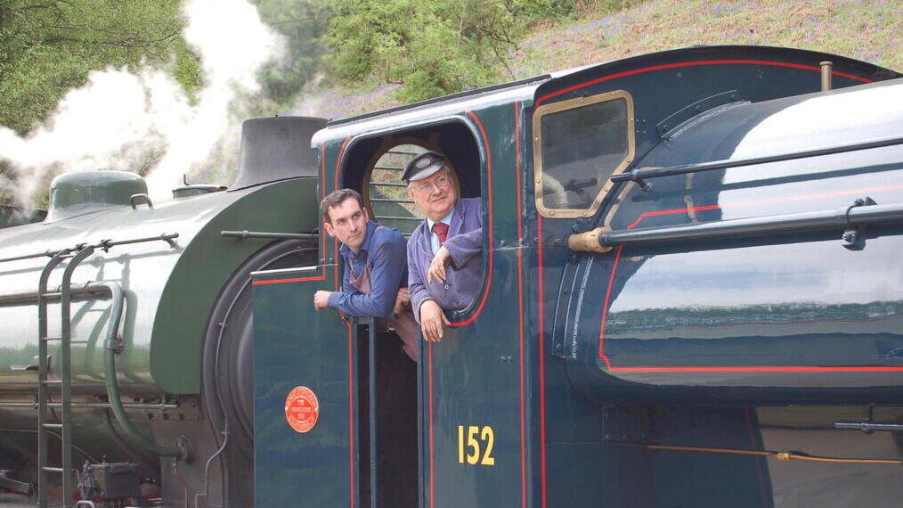 Two men wearing overalls standing in the cab of a vintage steam train.