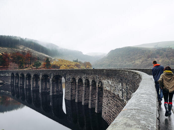 Two people walking on an arched bridge spanning a lake, with a valley in the background