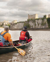 Zwei Menschen rudern auf einem Fluss in Richtung Chepstow Castle