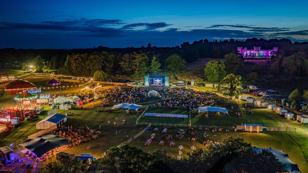 A large outdoor concert with a large screen with crowds and many tents and fairground rides.