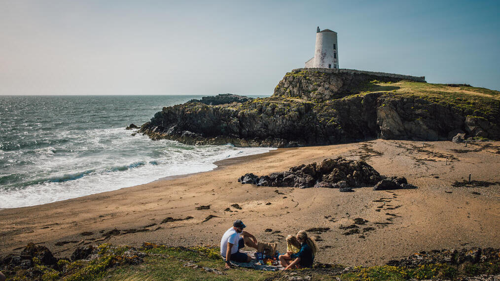Une jeune famille pique-niquant sur une plage avec un phare en arrière-plan.