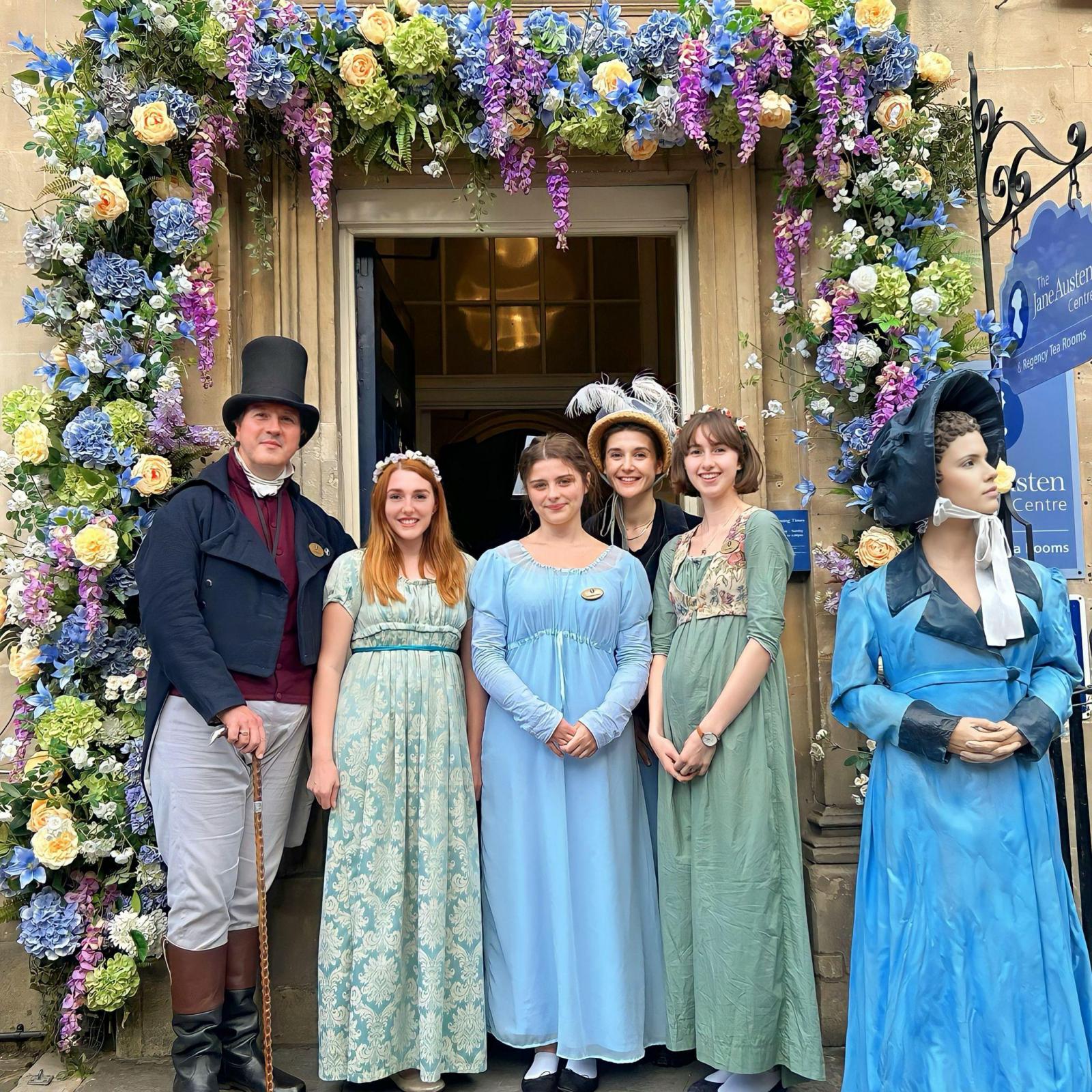 A group of people posing in period Victorian costume under a floral archway.