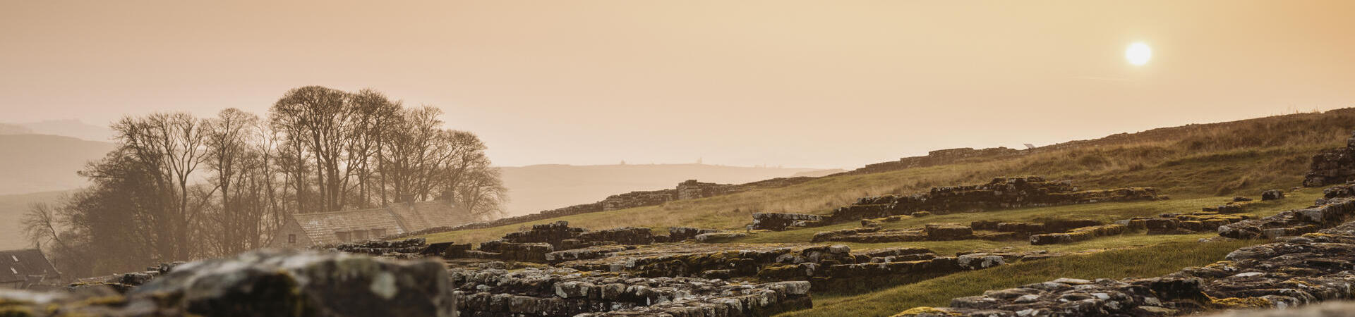 Ancient stone ruins on a grassy landscape at sunset, with distant trees and soft orange sky in the background.