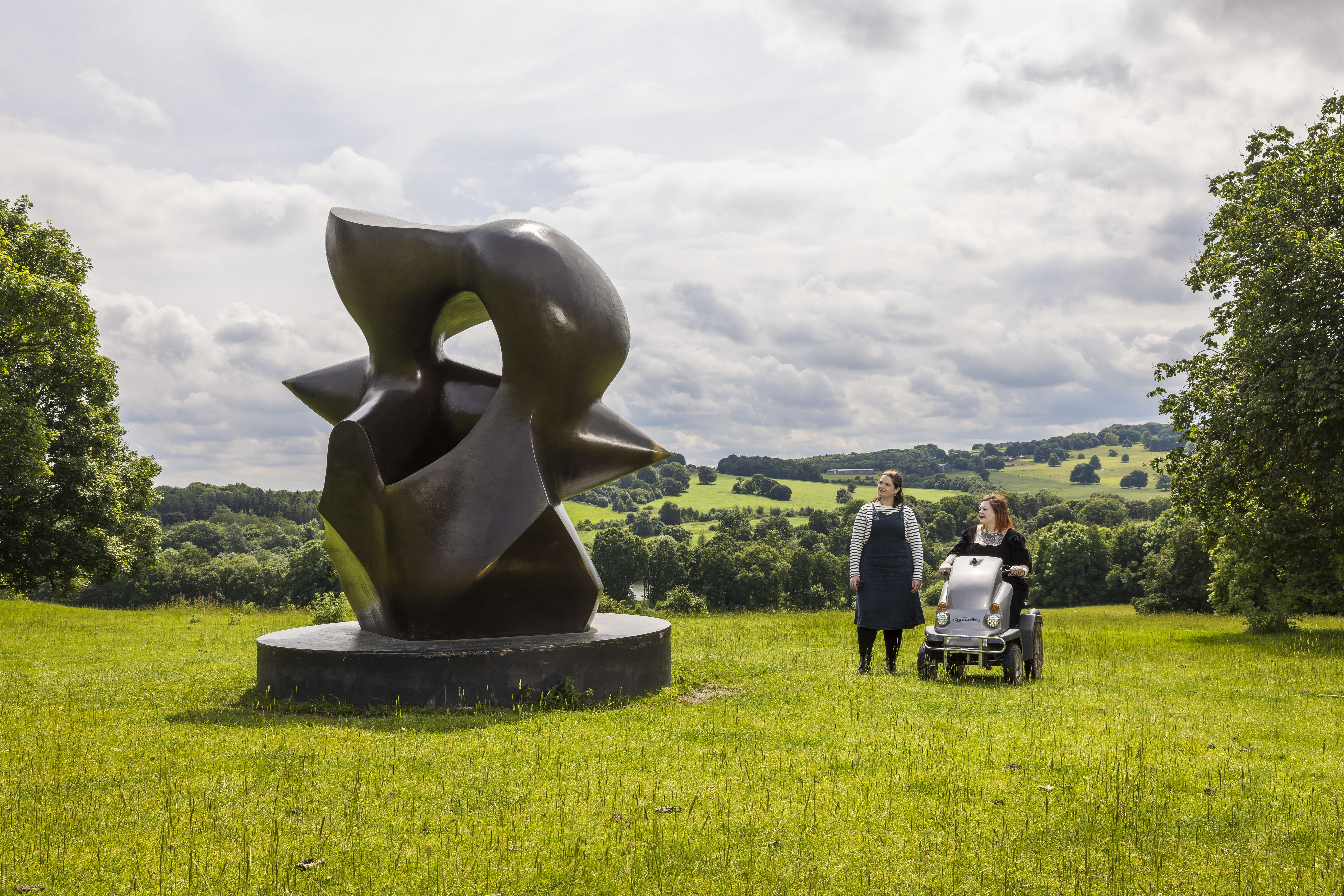 Two women look at a large sculpture set in green gardens with landscape beyond