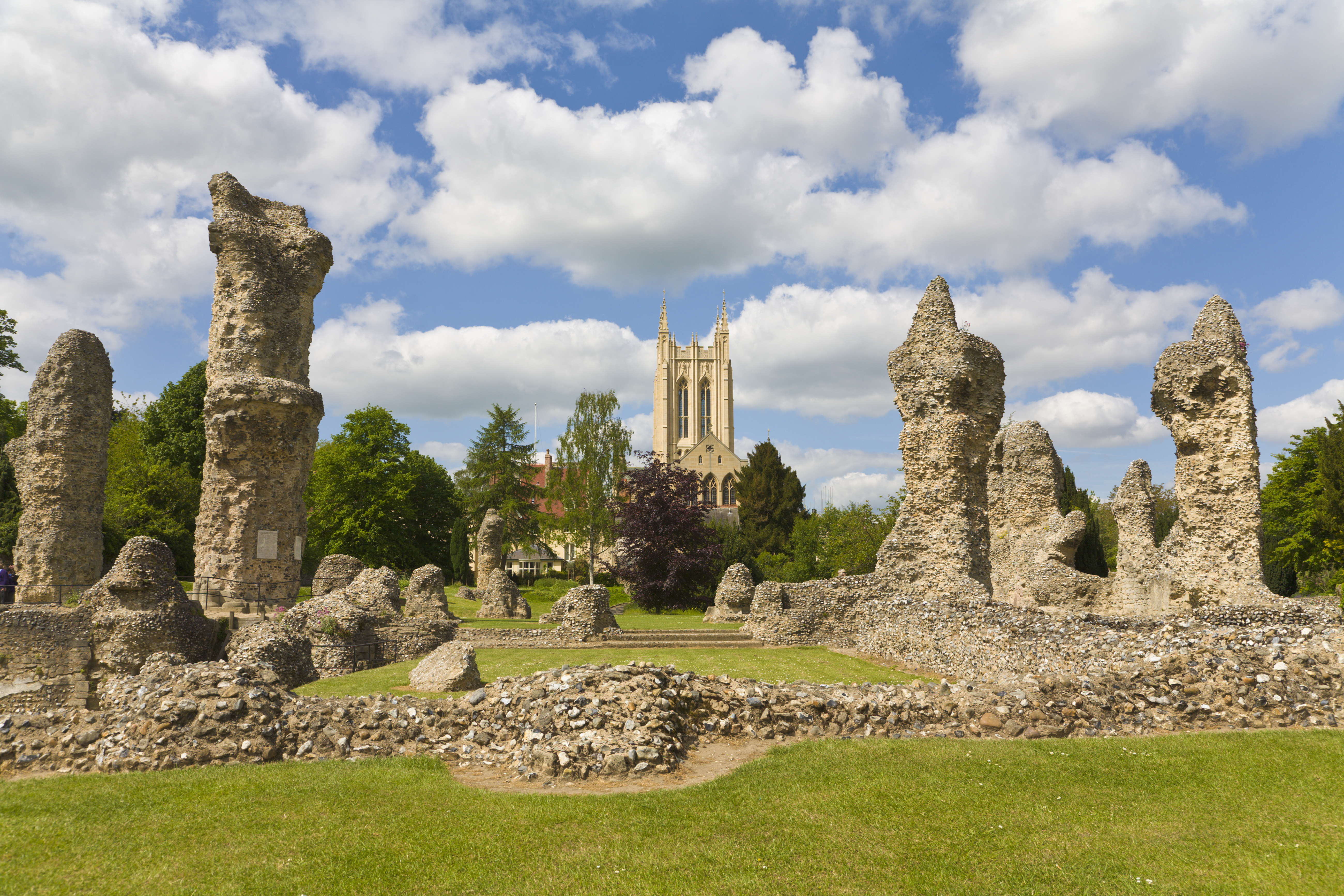 Ruinen einer Abtei mit einer Kathedrale im Hintergrund, blauer Himmel mit Wolken und Wiese ringsum.