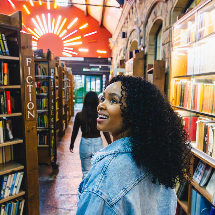 Two people walk through a vibrant, well-lit bookstore with shelves of books, stone walls, and neon lights overhead.