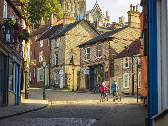 Two cyclists walking uphill with bikes in Lincoln, with Lincoln Castle in the background