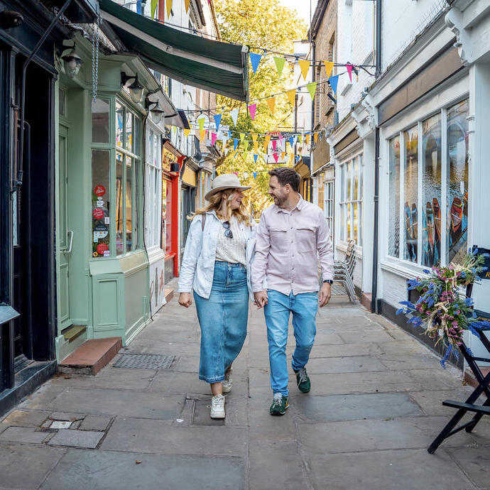 A couple walking and holding hands down a charming narrow street with colorful bunting overhead and shopfronts on either side.