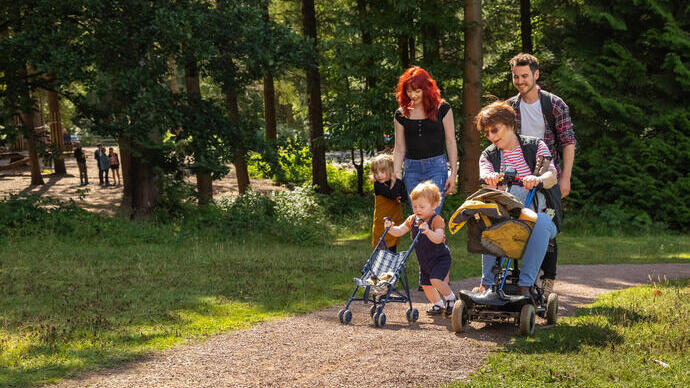 A family enjoying a walk together in a forest, with children playing and an adult using a mobility scooter on the path.