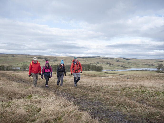 Four people hiking on grassy hills with patchwork fields and a lake in the background under a cloudy sky.