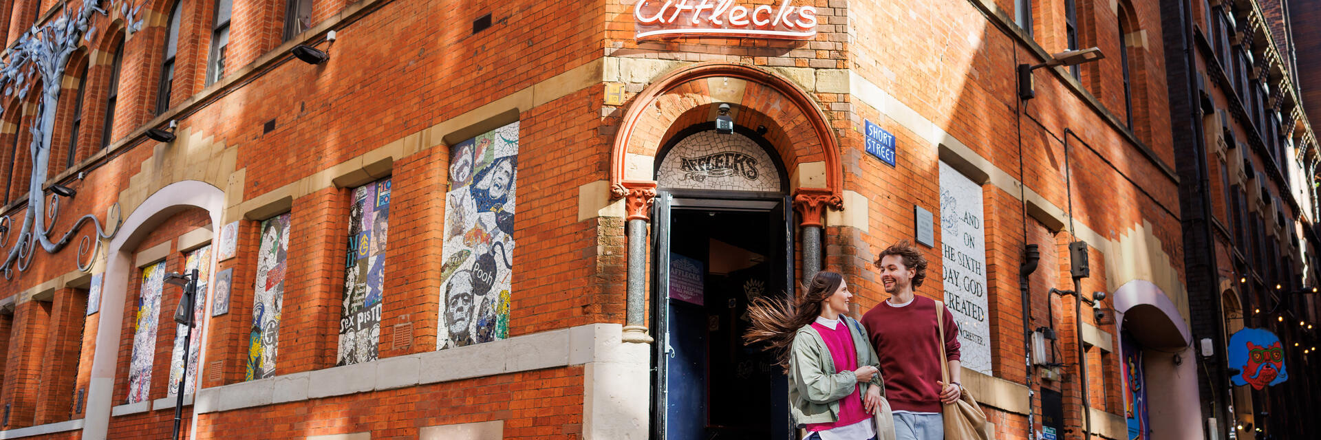 Two people walk outside Afflecks, a historic red-brick building in Manchester, with street art and urban scenery visible.