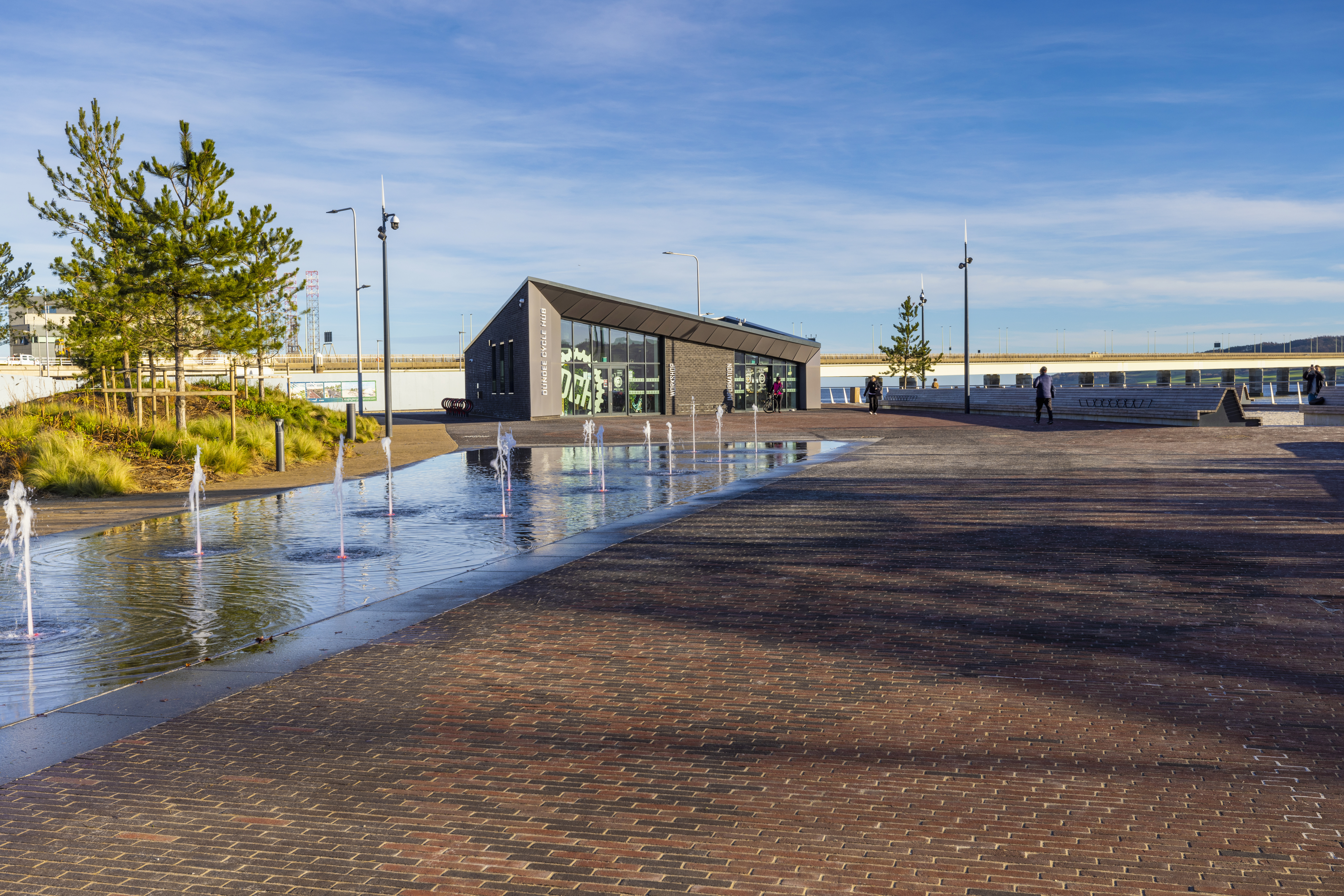 Modern waterside plaza with small fountains, a triangular building, landscaped greenery, and people walking under a clear blue sky.