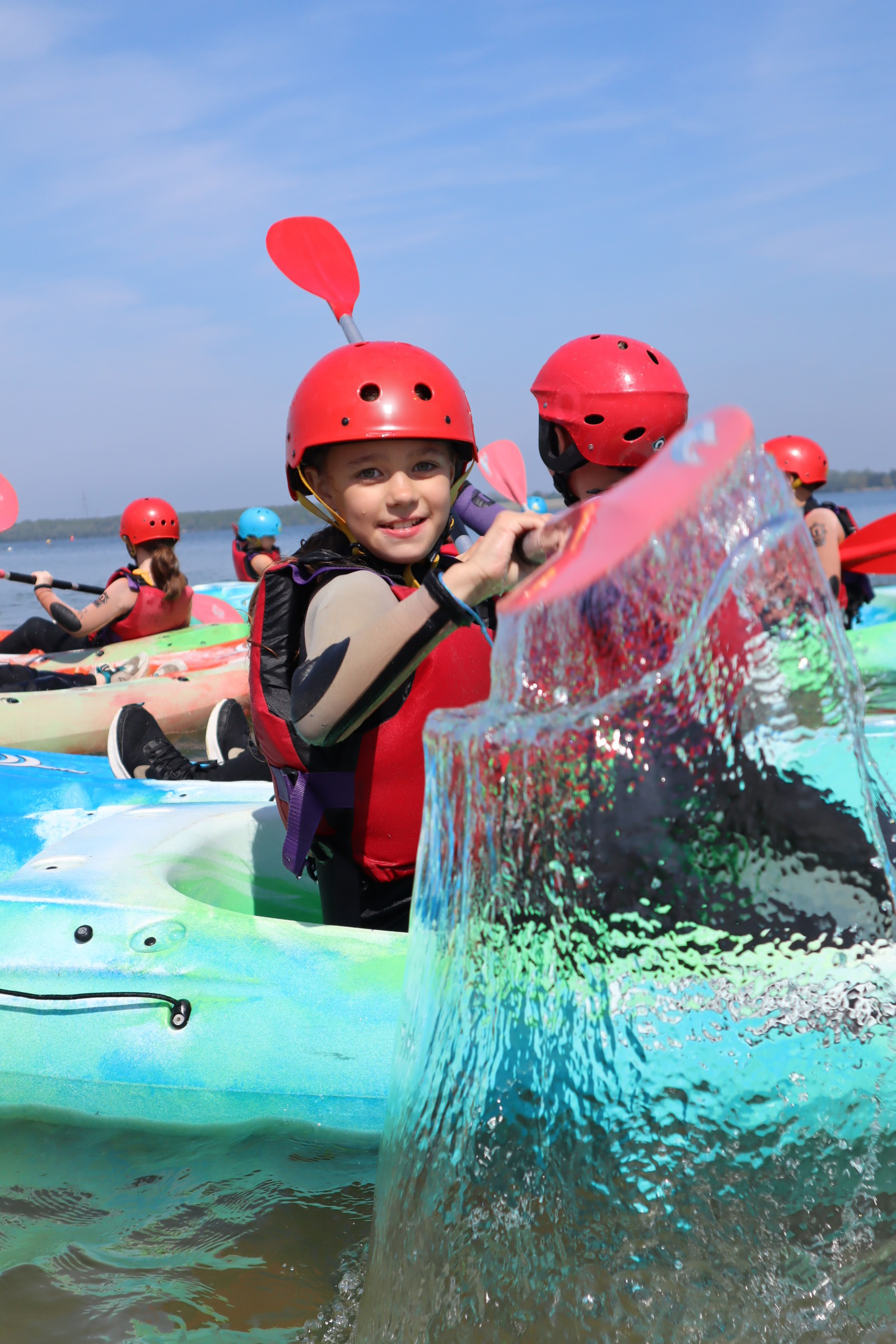 Children in helmets and life jackets kayaking on a lake under a clear blue sky, water splashing in foreground.