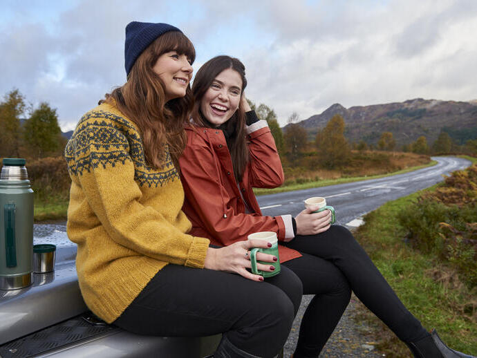 Zwei Personen sitzen auf einer Motorhaube, trinken aus Tassen in ländlicher Umgebung, mit Thermoskanne daneben und einer kurvigen Straße im Hintergrund.