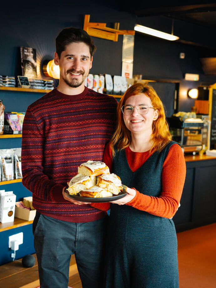 A woman and a man hold a plate of cinnamon buns in a cafe