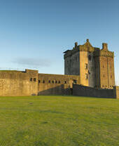 Historic stone castle with fortified walls on green grass, beside a body of water under a clear blue sky at sunset.
