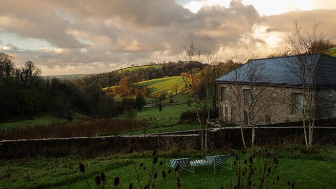 Stone house with slate roof beside green hills, trees, and dramatic cloudy sky. Two garden benches on grass in foreground.
