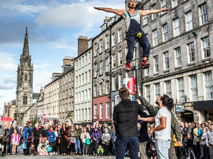 Man balancing high up on a unicycle and being held by helpers as a crowd stand watching in the street