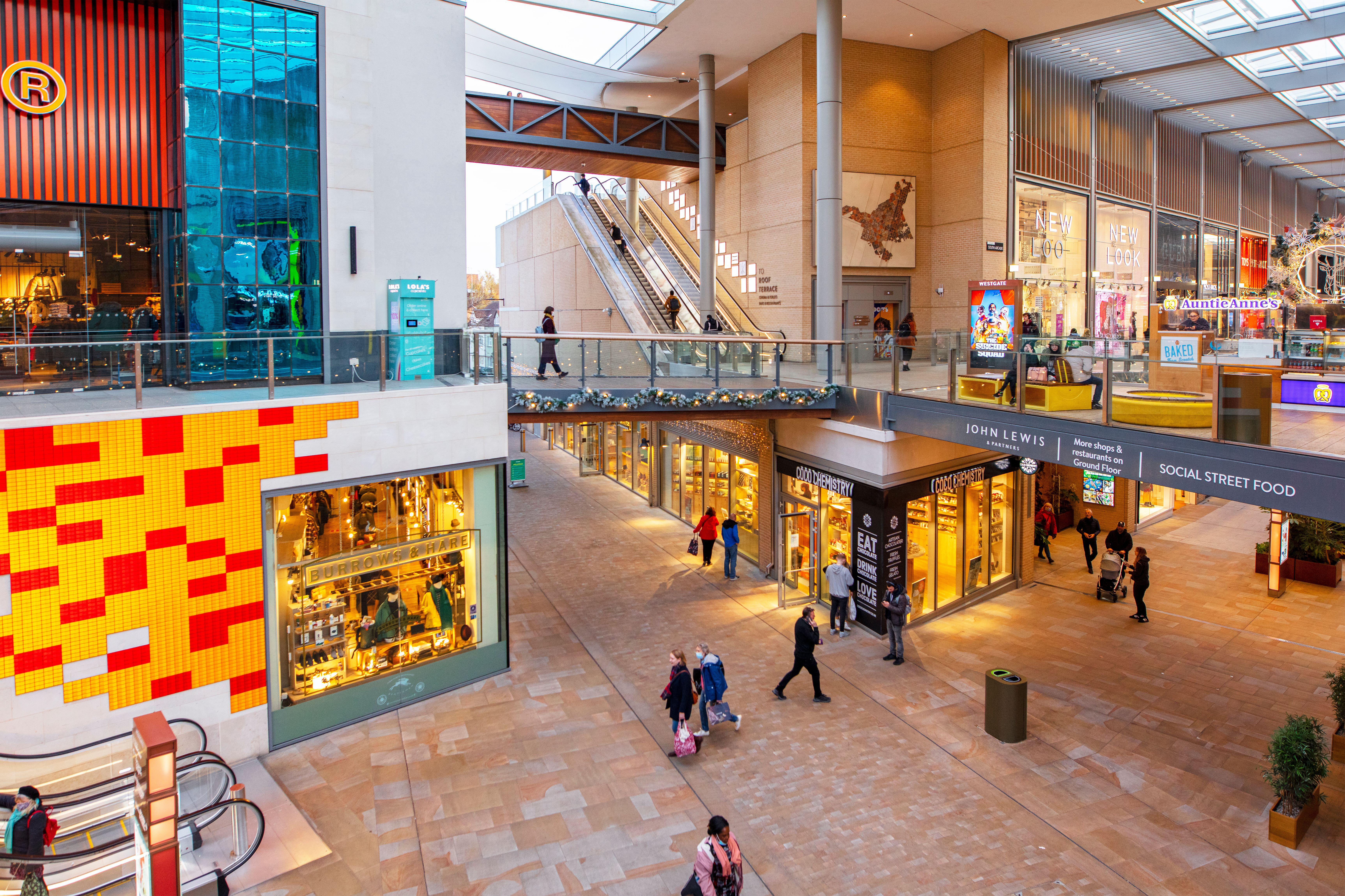 Modern indoor shopping mall with shops, escalators, colorful decor, glass railings, and people walking or shopping.