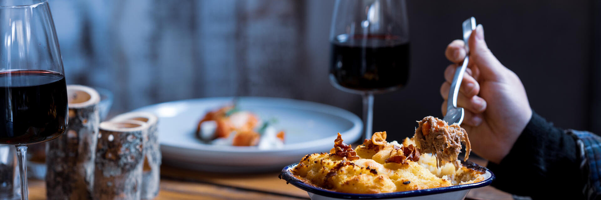 View of arm eating a mashed potatoes topped dish with two glasses of red wine and another dish of food in soft focus in the background 