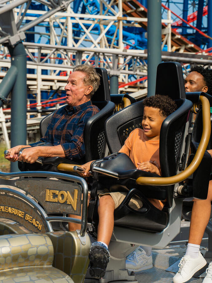 A group of people of different ages enjoying a roller coaster ride at Blackpool Pleasure Beach with bright coaster tracks in the background.