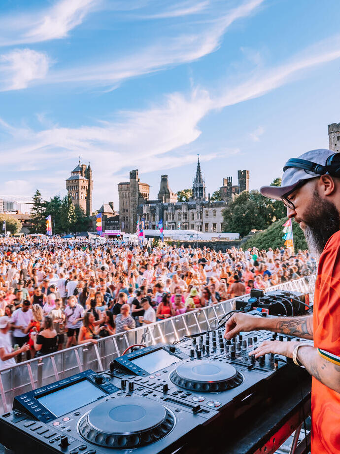 DJ spielt vor großem Publikum auf einem Musikfestival im Freien, mit einer Burg im Hintergrund und blauem Himmel.