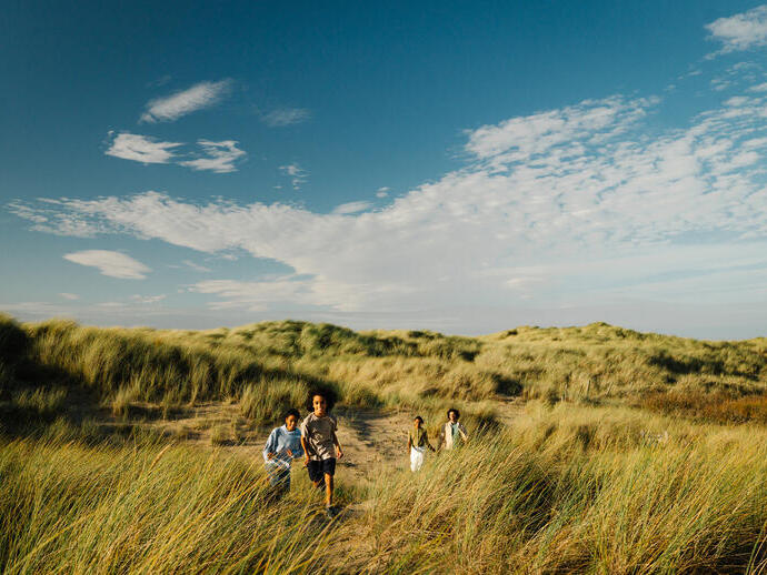 Children running through grassy sand dunes under a bright blue sky with scattered clouds.