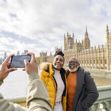 A man photographing mature couple with a large river and iconic buildings in the background.