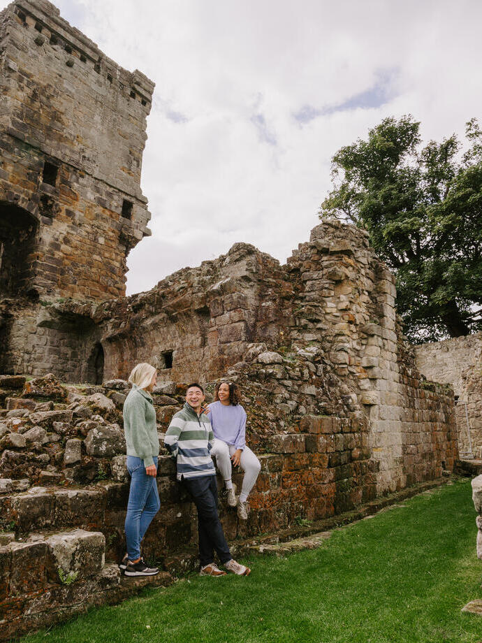 Two women and a man sitting on the ruins of a large castle and garden area.