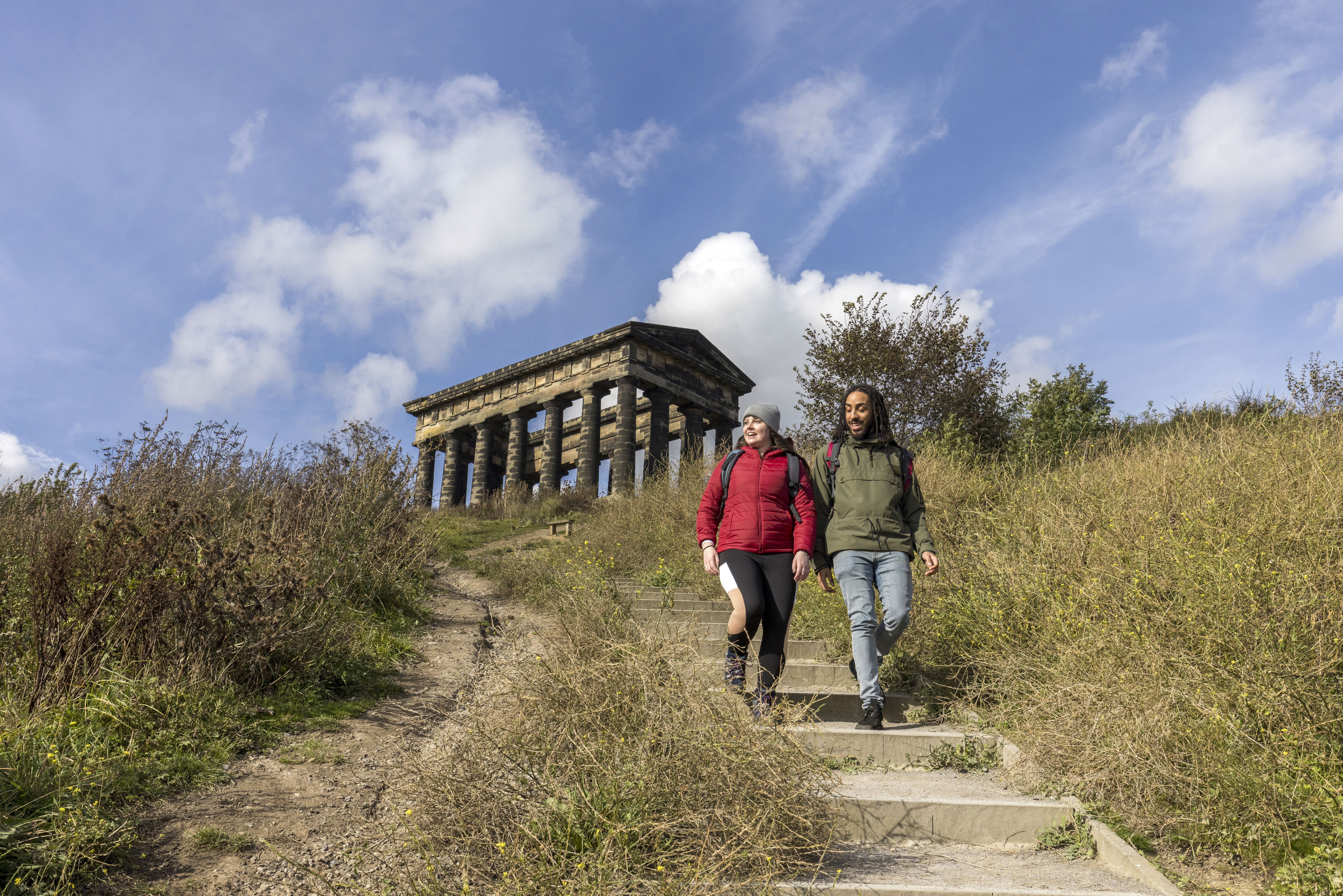 Two people wearing outdoor clothing, hiking past a stone monument, surrounded by greenery and blue skies.