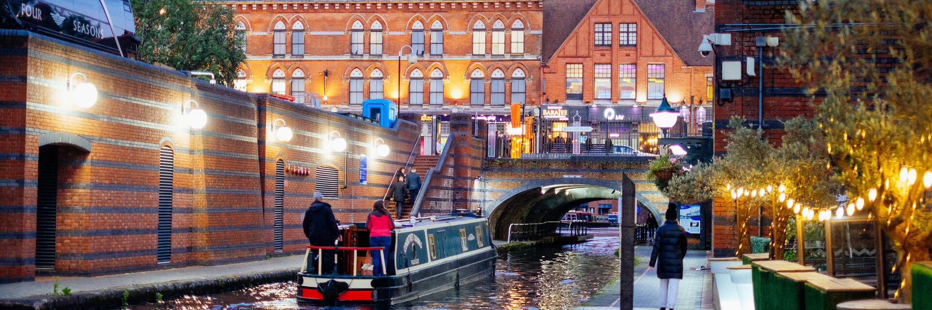 Flussboot, das in der Abenddämmerung einen Kanal hinunterfährt, während Menschen auf einem parallel verlaufenden Weg spazieren gehen.