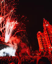 Crowds of people watching a band at a music festival as fireworks explode, in front of the Liver Building.