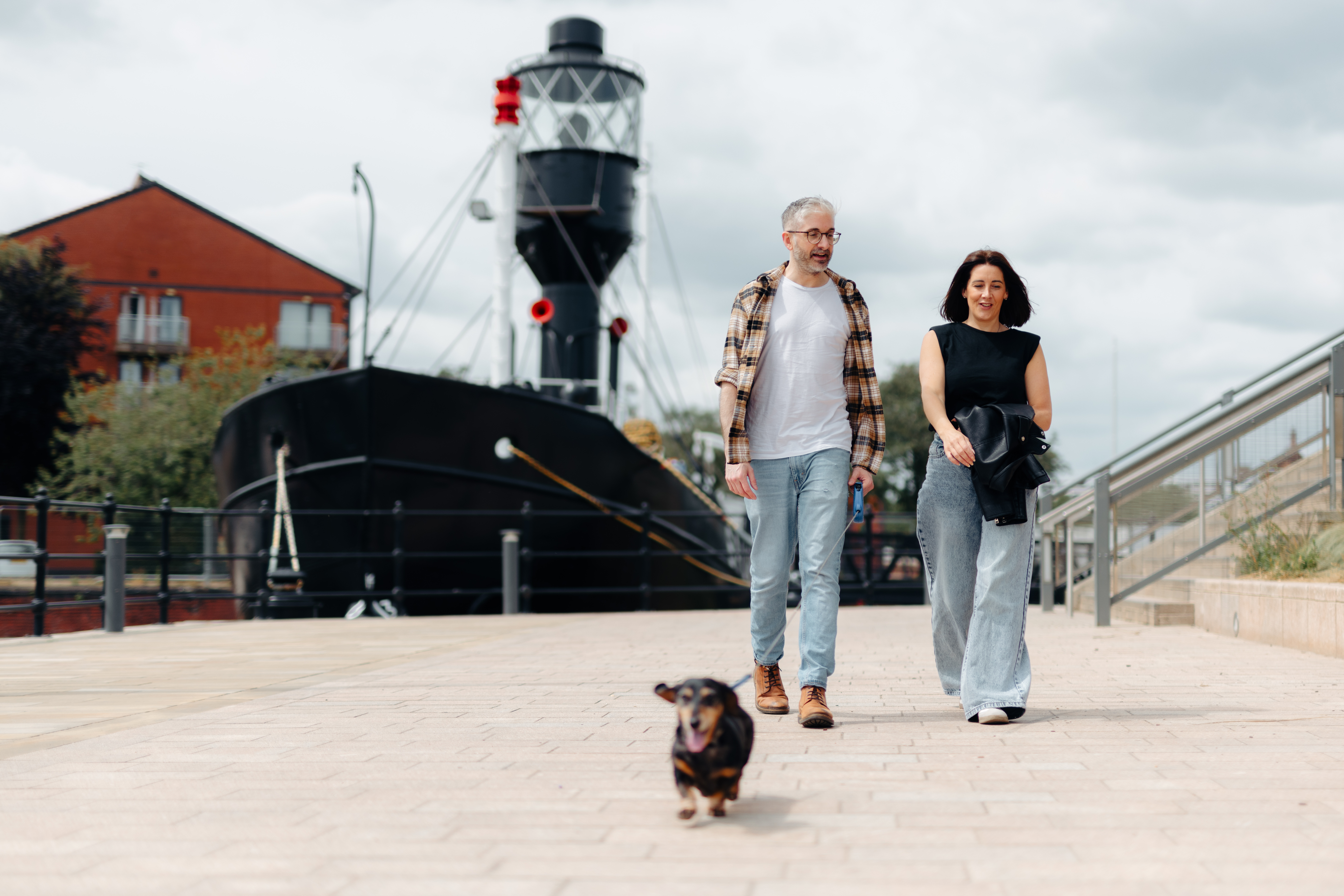 Two people walking a small dog along a paved waterfront with a historic ship and residential buildings in the background.