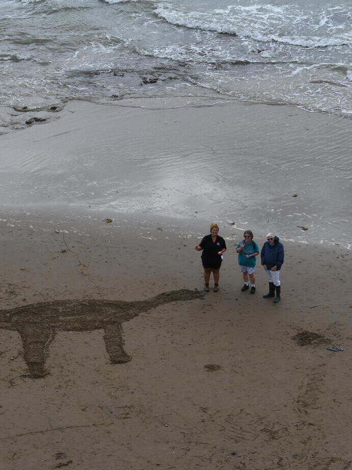 People on a beach with a dinosaur carved into the sand