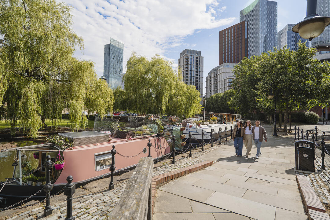 Canal with moored narrowboats, leafy trees, and modern city buildings in the background. Three people walk along the paved path.