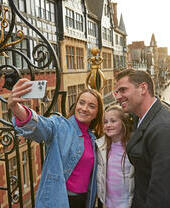 A family taking a selfie on a historic street with ornate ironwork and timber-framed buildings in the background.