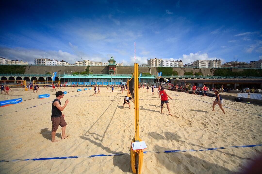 People playing beach volleyball on a sand court with large buildings and a blue sky in the background.