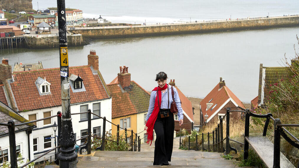 Person walking up stone steps with red-roofed houses below, seaside harbour, and sea walls in the background. Coastal village scenery.