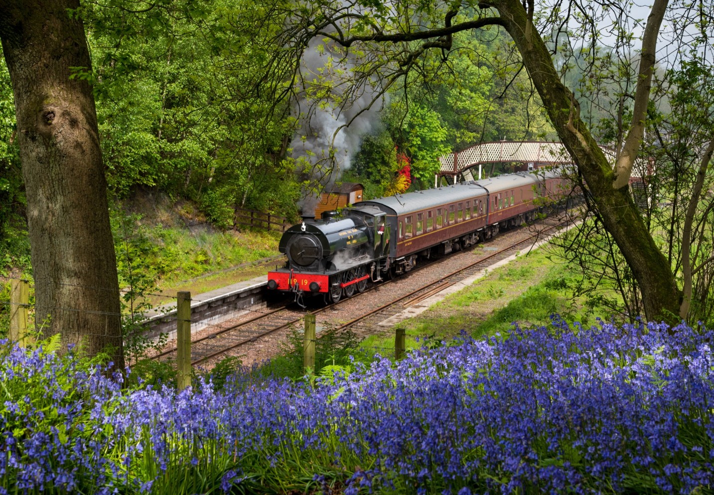 A steam train with lots of steam coming out, pulling at least three vintage carriages, gets ready to depart a small traditional railway station.