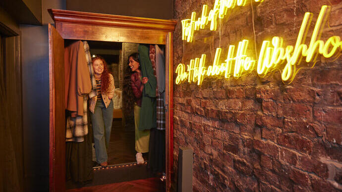 Two people emerge from a wardrobe into a room with exposed brick and neon sign, evoking a secret passage or escape room theme.