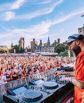 DJ spielt vor großem Publikum auf einem Musikfestival im Freien, mit einer Burg im Hintergrund und blauem Himmel.