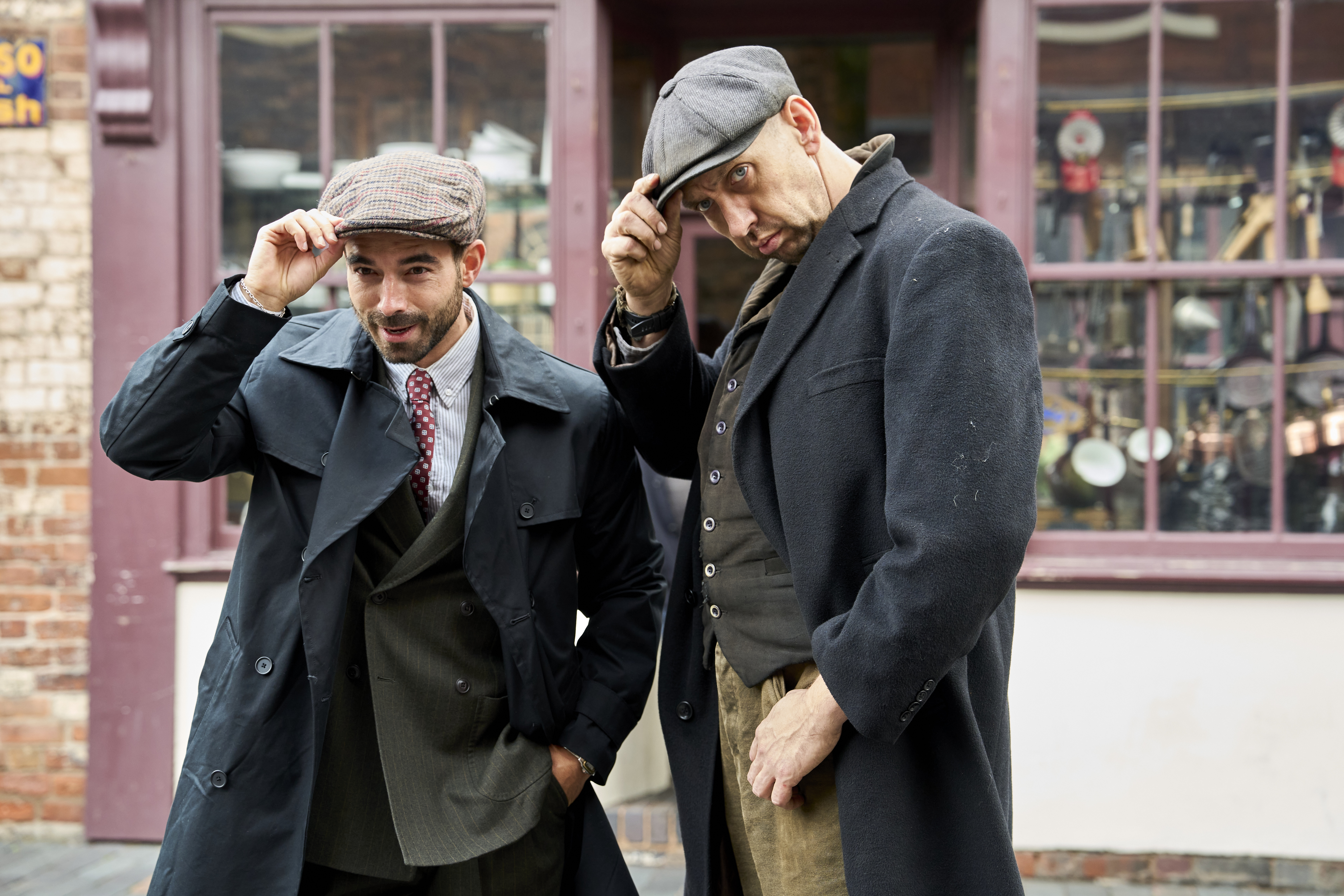 Two men in vintage clothing and flat caps pose outside a shop with a display window and brick wall backdrop.