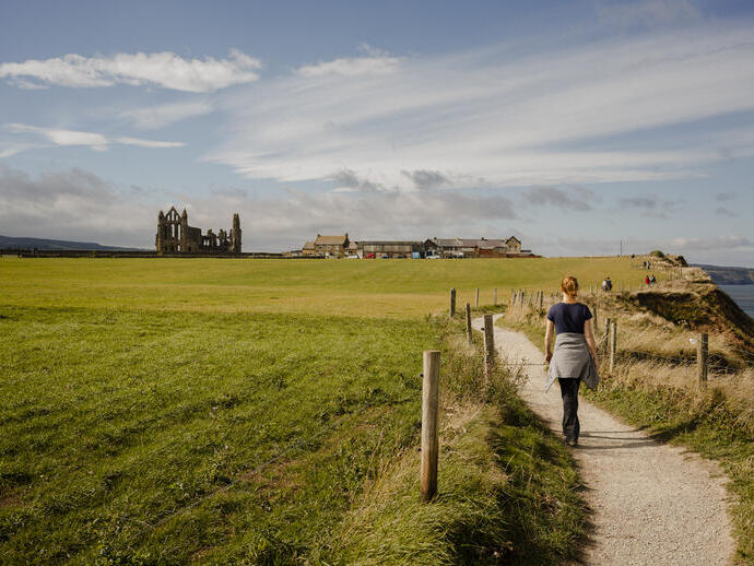 Woman walking along a coastal path towards a ruined abbey